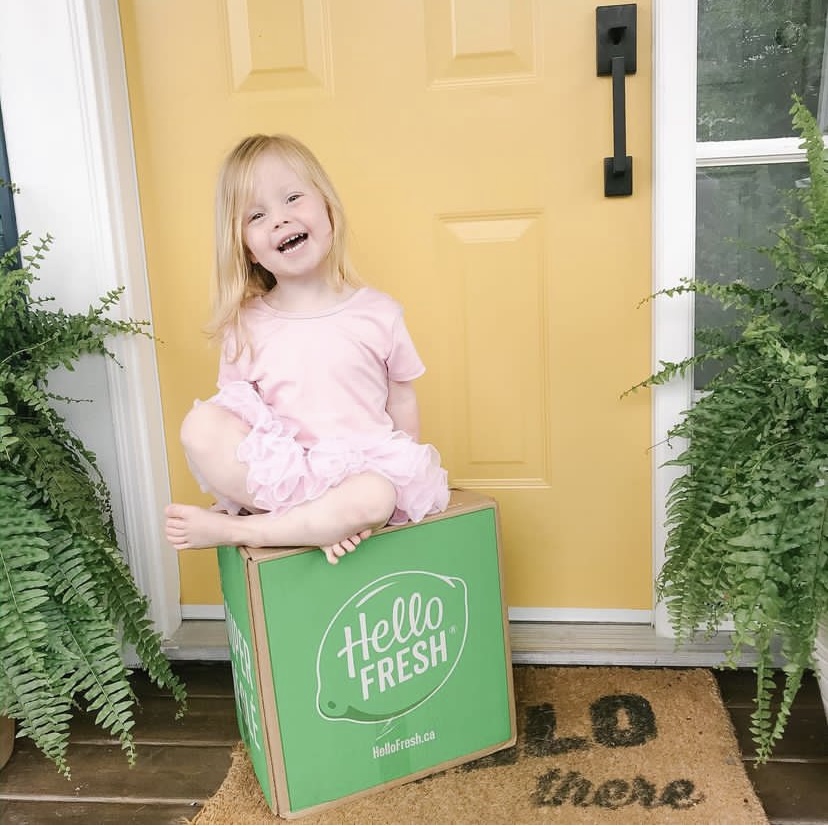 Iyla Coughlin sits on top of a Hello Fresh food delivery box on the front door step with a big smile on her face